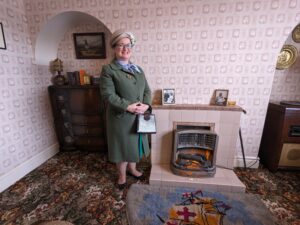 A lady in 1950s clothes stands in front of a fireplace. The room is decorated in a 1950s style.